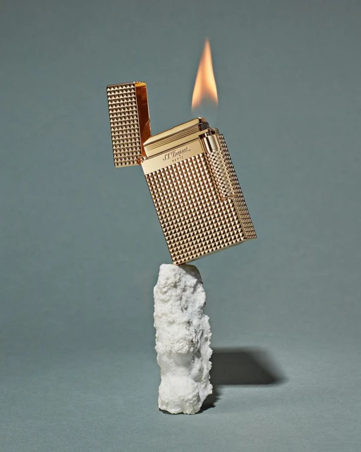 Gold textured lighter with flame on a white rock against a blue background, promoting luxury and style in a studio product shot.
