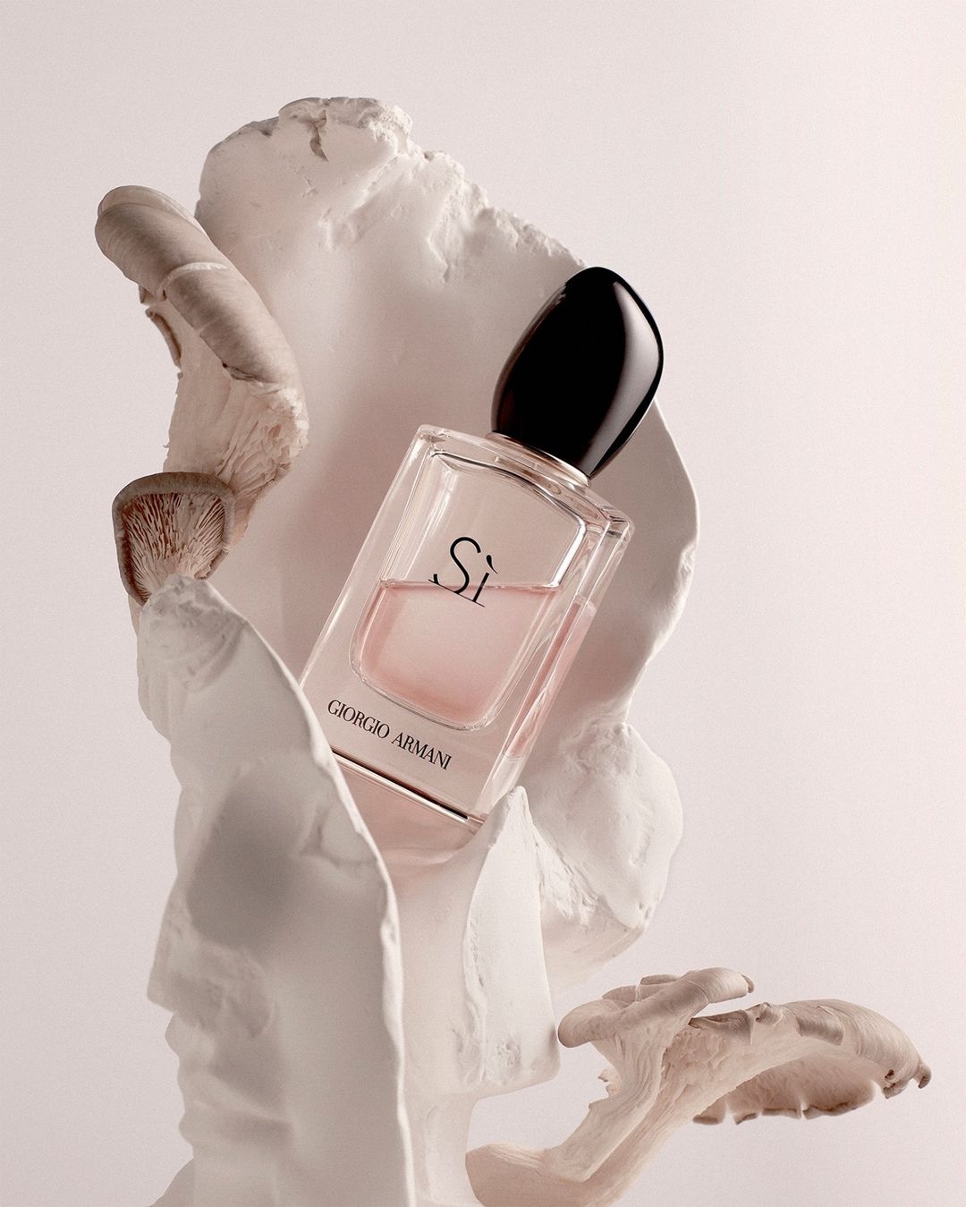 Giorgio Armani Si perfume bottle amidst a sculpted white bust and cream-colored mushrooms, set against a soft, light background.