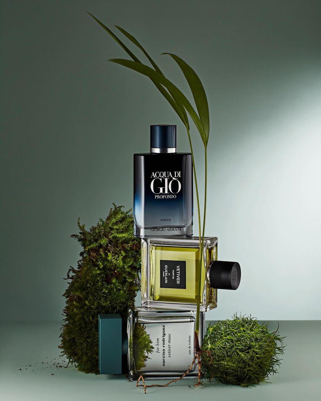 Three stacked fragrance bottles surrounded by green moss and botanical sprigs against a gradient green backdrop in a studio setting.