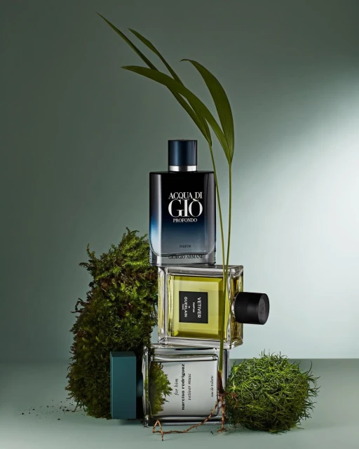 Three stacked fragrance bottles surrounded by green moss and botanical sprigs against a gradient green backdrop in a studio setting.
