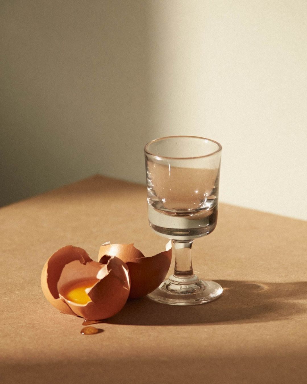 Broken eggshells and a clear footed glass are displayed on a tan surface with a creamy background, illuminated by soft light.