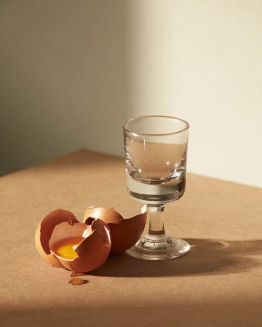 Broken eggshells and a clear footed glass are displayed on a tan surface with a creamy background, illuminated by soft light.