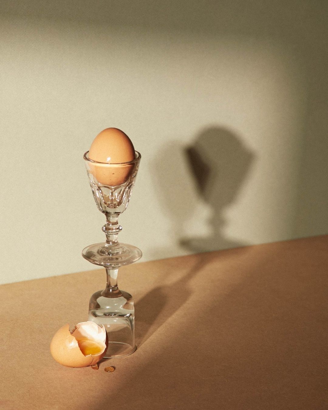 Still life shot of a brown egg in a stemmed glass atop an upside-down wine glass, with an eggshell on a beige surface.