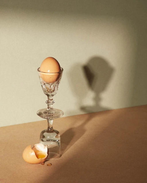 Still life shot of a brown egg in a stemmed glass atop an upside-down wine glass, with an eggshell on a beige surface.