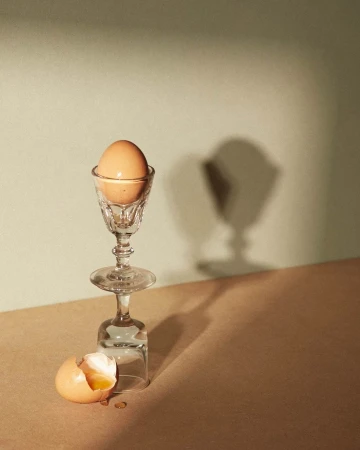 Still life shot of a brown egg in a stemmed glass atop an upside-down wine glass, with an eggshell on a beige surface.