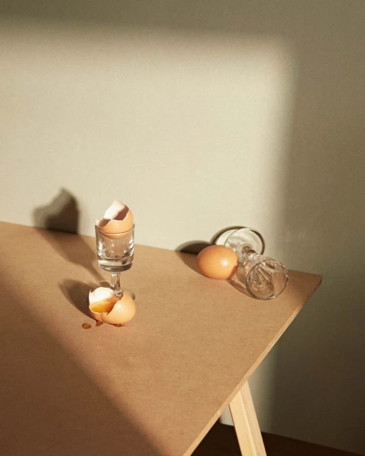 Still life featuring cracked eggs in glass egg cups on a wooden table, bathed in warm, angled sunlight creating shadows on a pale green wall.