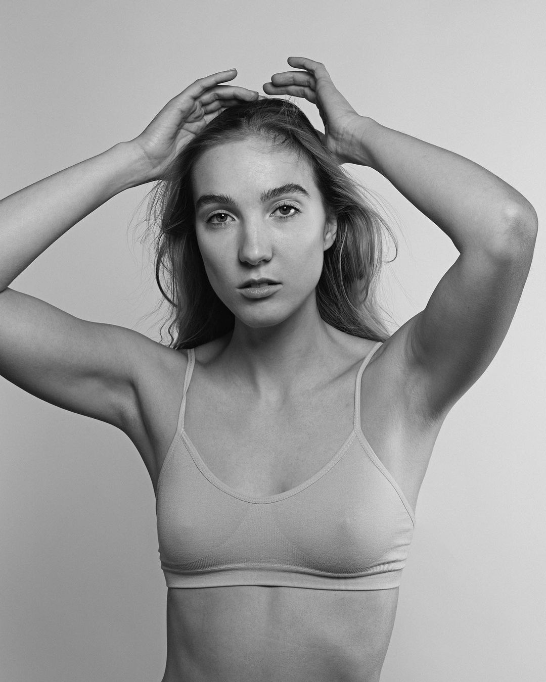 Monochrome portrait of a young woman with wavy hair and a neutral facial expression, hands on her head, wearing a bra top in a studio.