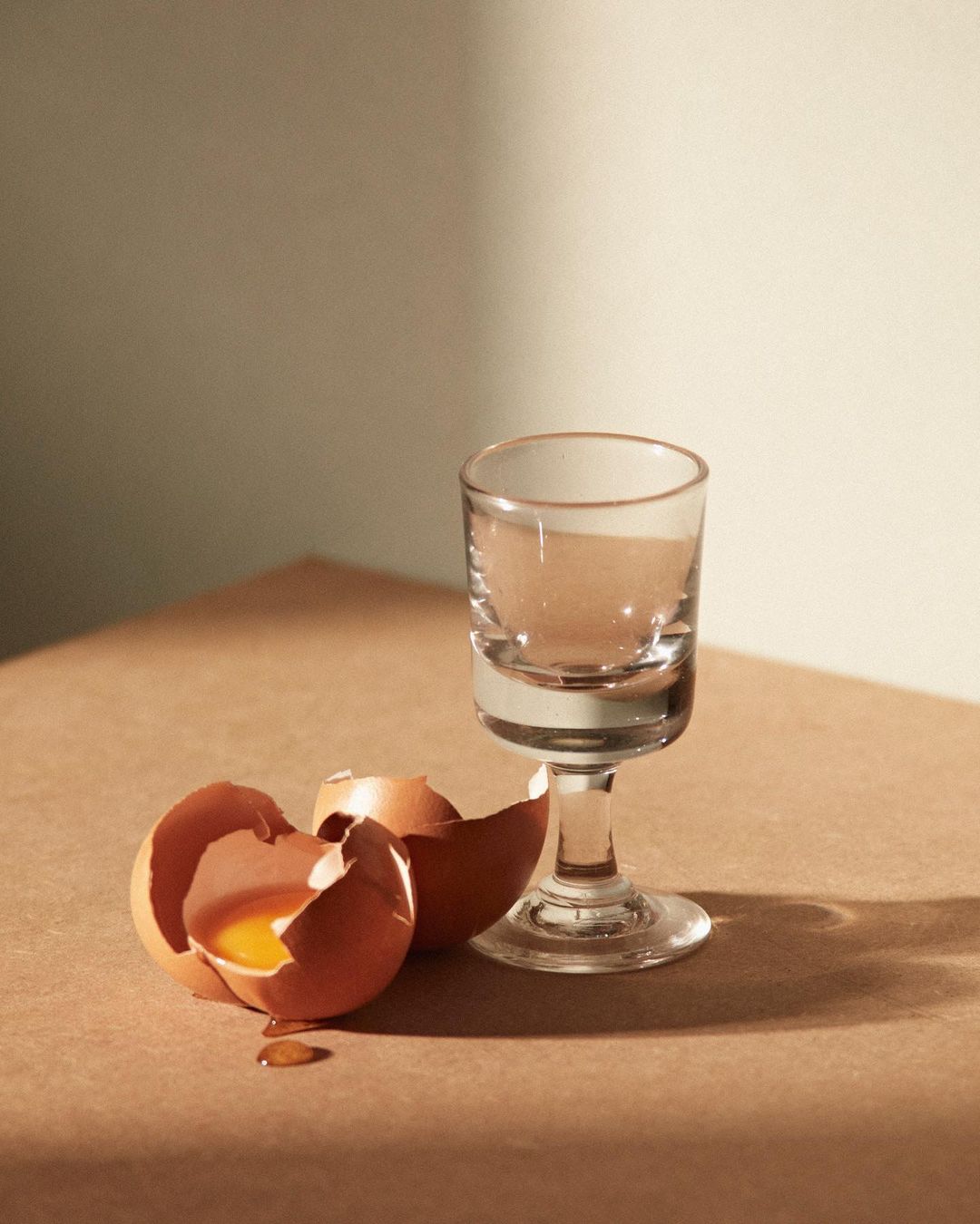 Still life shot of an egg shell with yolk, and a stemmed glass on a tan surface with a light beige background in studio light.