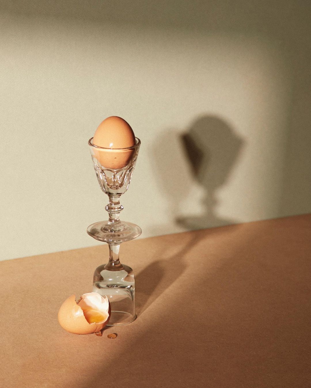 Still life of a cracked egg with flowing yolk, clear crystal glasses, and soft studio lighting on a neutral background.