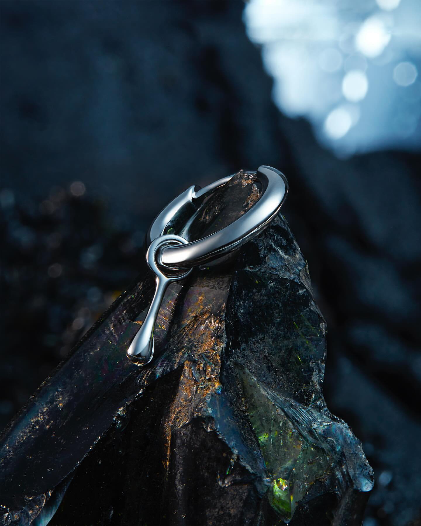 Macro shot of a silver metal earring pendant resting on a textured black rock, set against a blurred blue and black background.