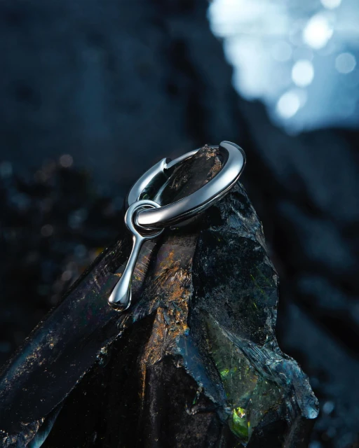 Macro shot of a silver metal earring pendant resting on a textured black rock, set against a blurred blue and black background.