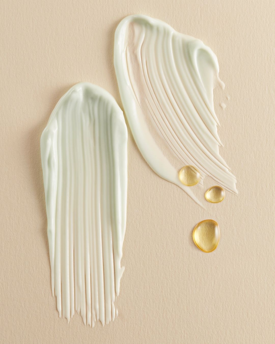 Swatches of light green cream product and drops of golden oil on a beige background, showcasing cosmetic textures for beauty advertisement.