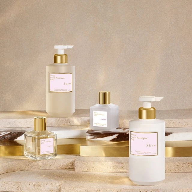 Four bottles of Maison Francis Kurkdjian à la rose hand soap and lotion displayed on marble and gold platforms against a beige backdrop.