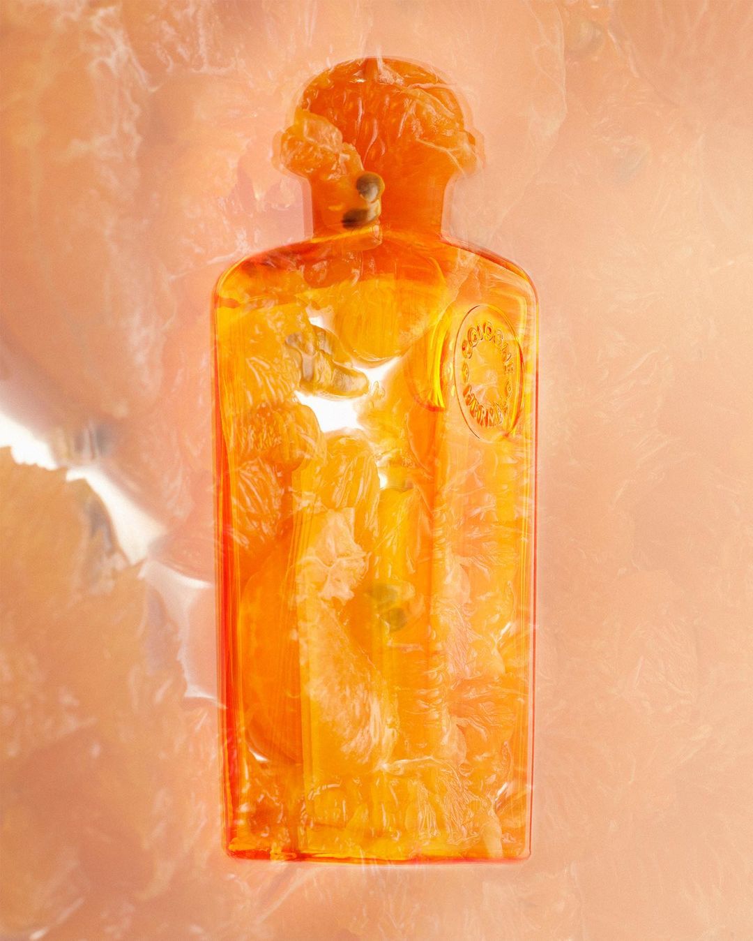 Conceptual still life of a translucent orange perfume bottle filled with orange slices, set against a blurred orange background.