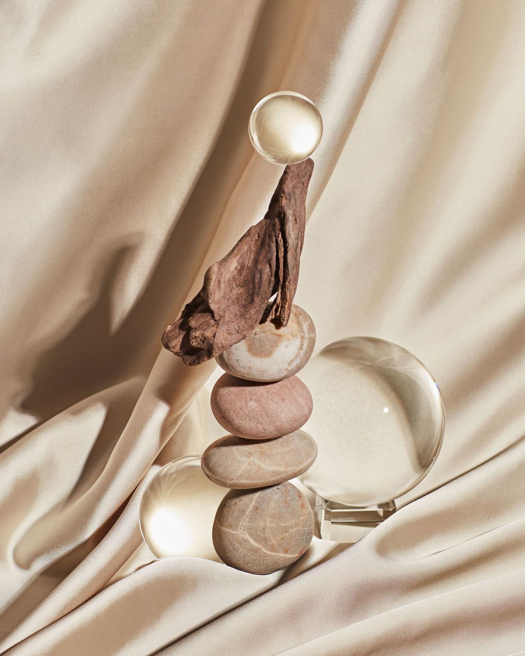 Still life featuring balanced stones, clear orbs, and a piece of wood, set against a smooth, draped beige fabric background.