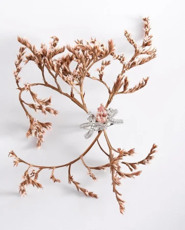 Elegant diamond ring with a pink gemstone is displayed on dried floral branches against a clean white backdrop, jewelry still life.