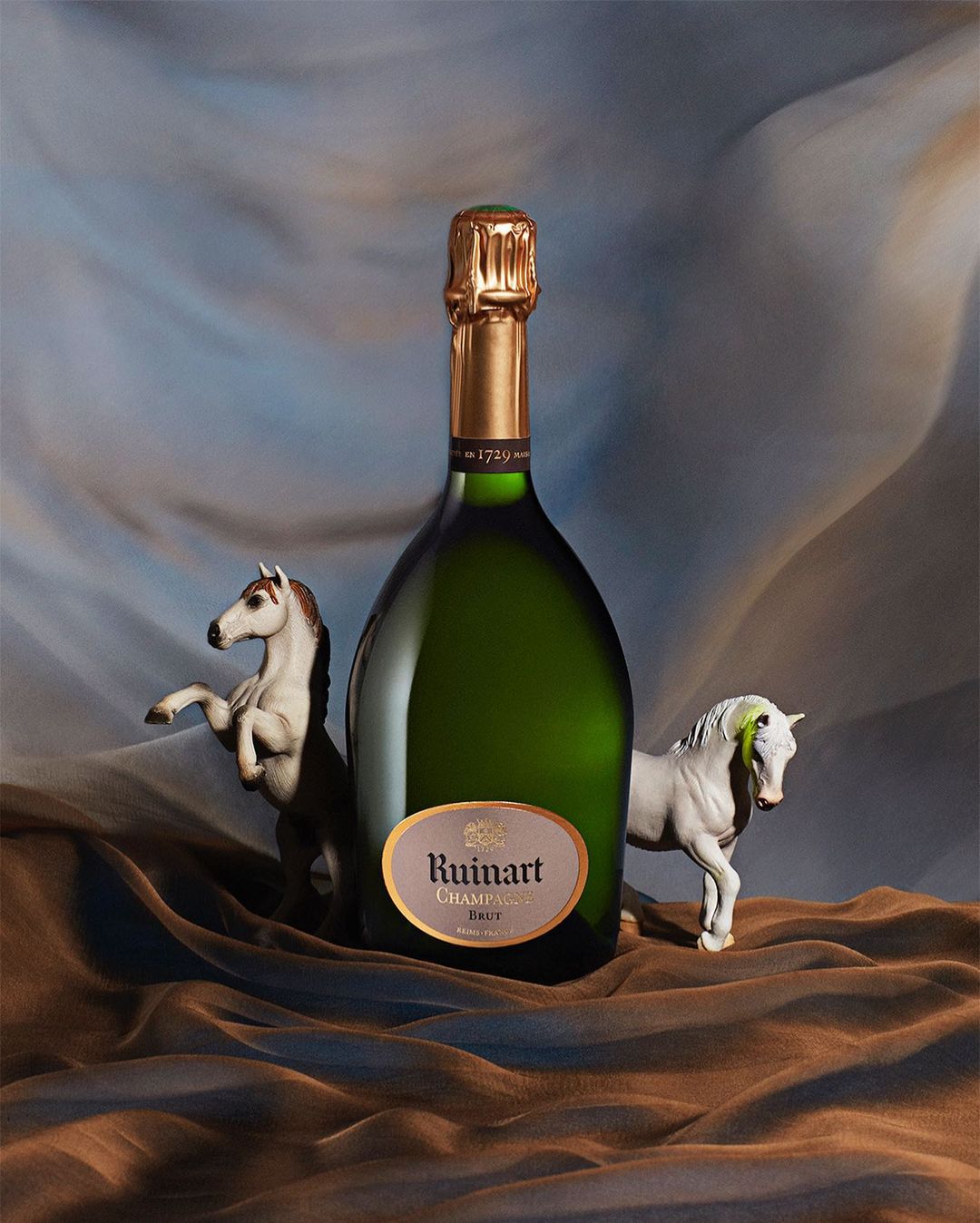 Studio shot of Ruinart champagne bottle with gold foil top, flanked by white toy horses on draped blue and brown fabric.