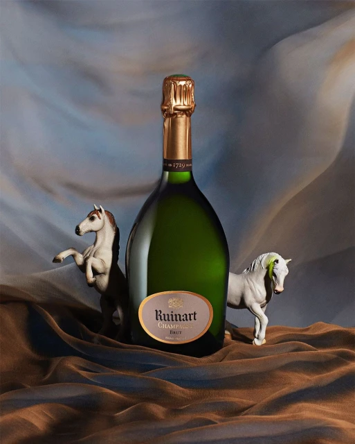 Studio shot of Ruinart champagne bottle with gold foil top, flanked by white toy horses on draped blue and brown fabric.
