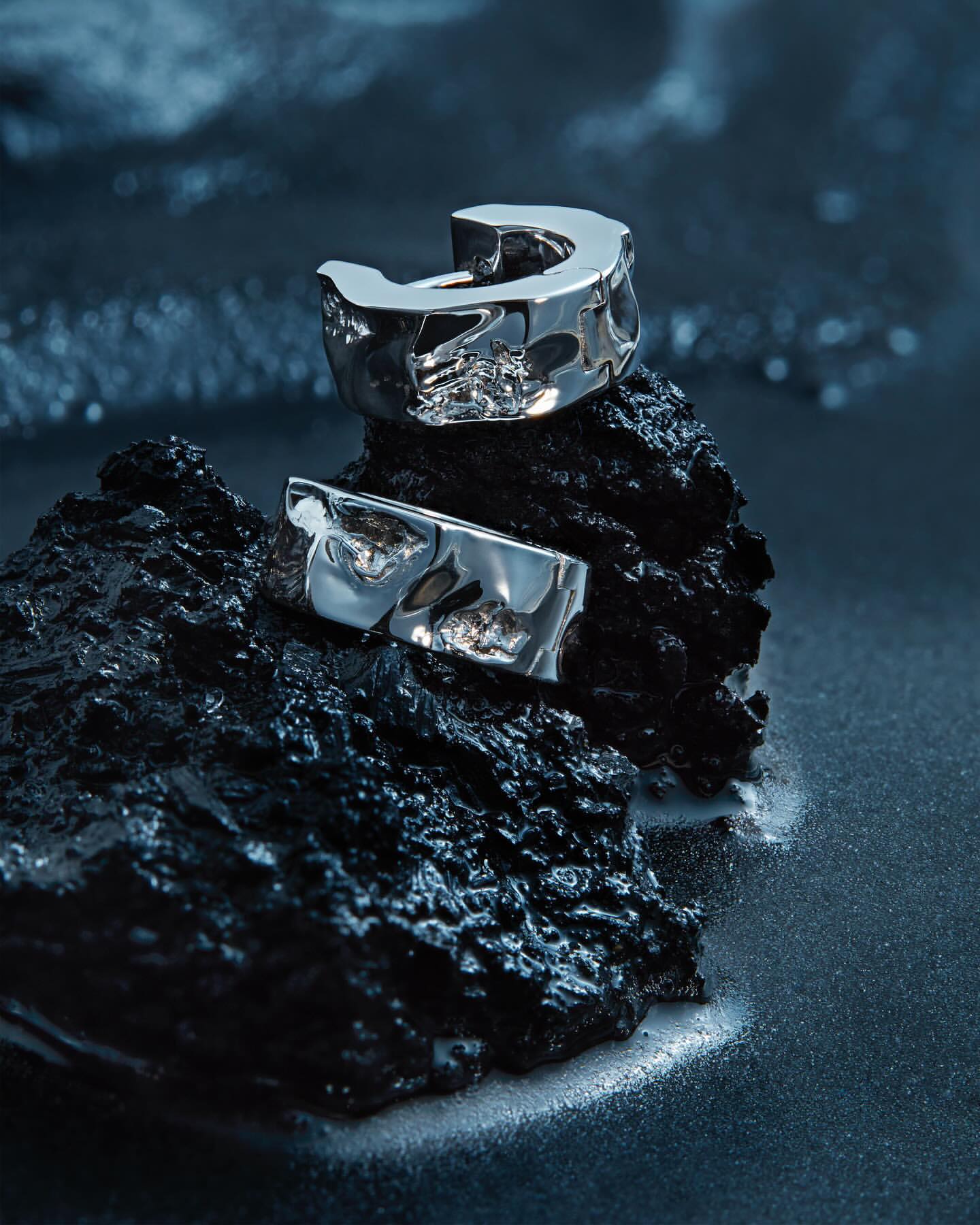 Close-up of two silver-toned earrings resting on a textured black rock against a dark blue shimmering background in a beauty shot.