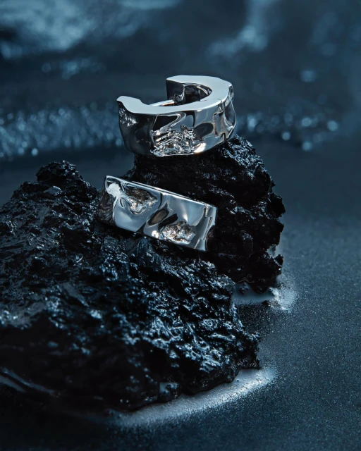 Close-up of two silver-toned earrings resting on a textured black rock against a dark blue shimmering background in a beauty shot.