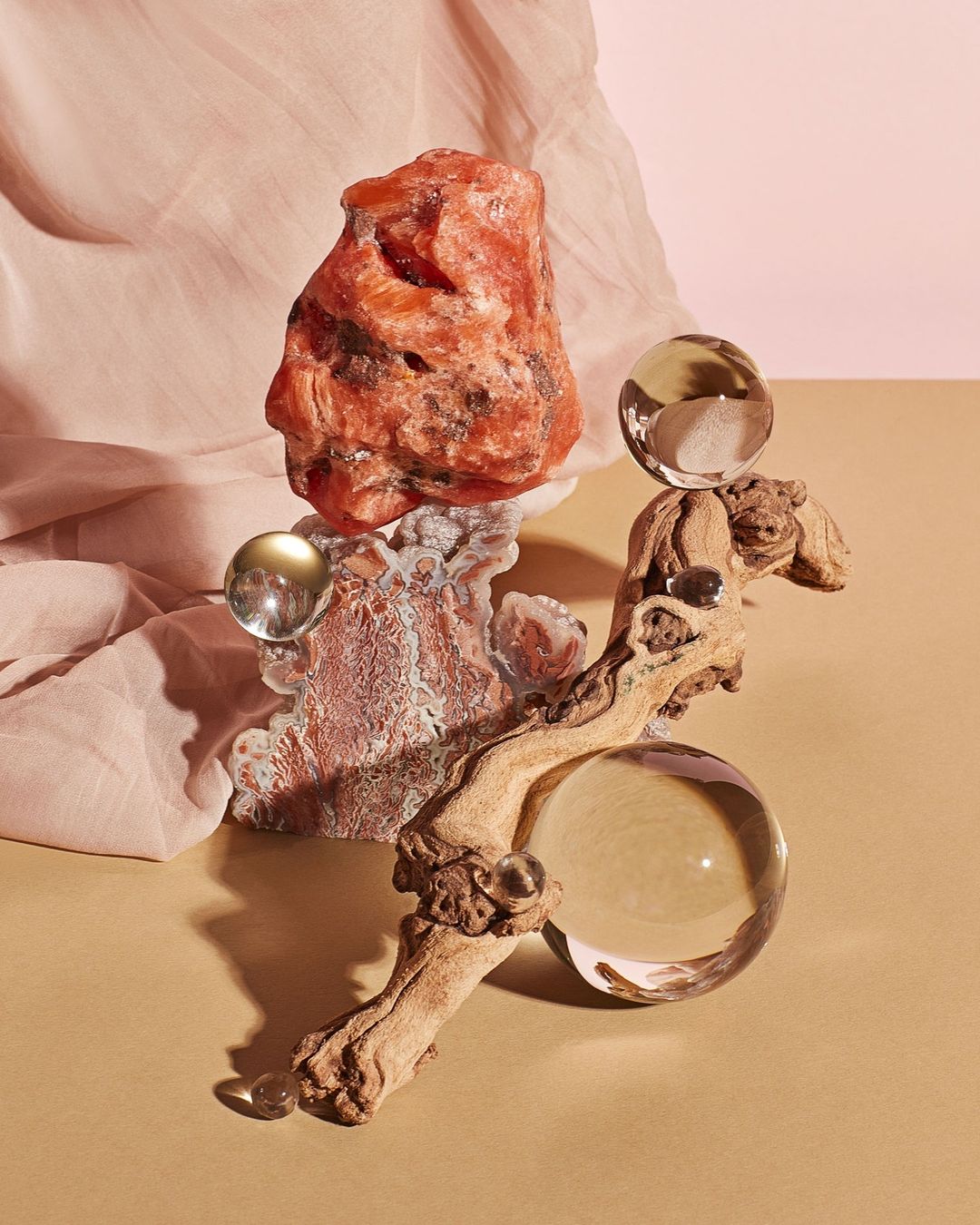 Still life photograph of a translucent orange rock balanced on a textured rock formation and piece of driftwood with clear glass spheres.
