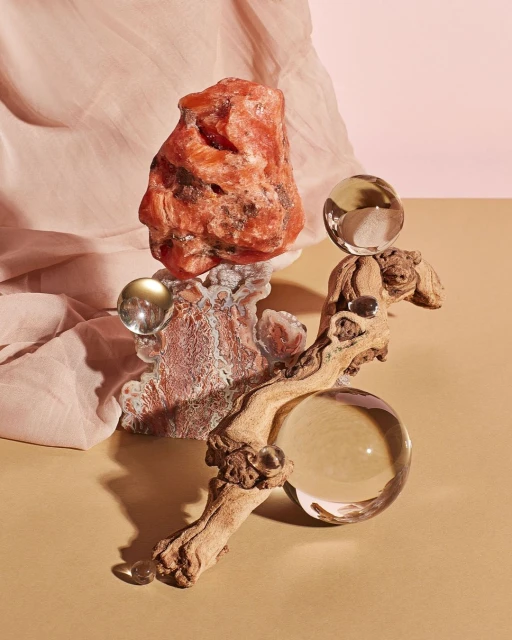 Still life photograph of a translucent orange rock balanced on a textured rock formation and piece of driftwood with clear glass spheres.