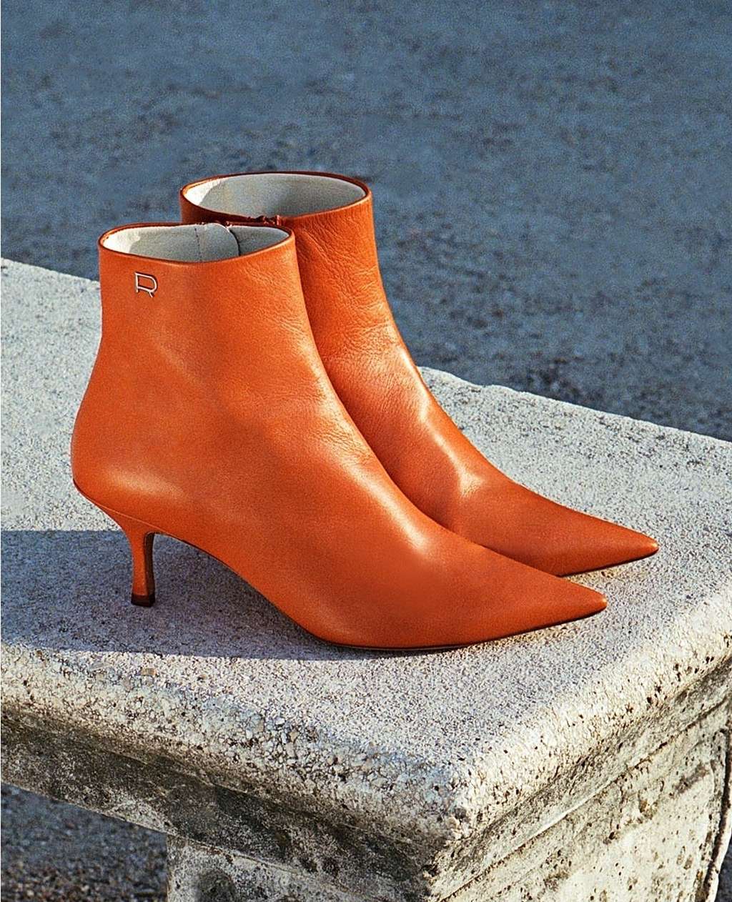 Pair of orange leather ankle boots with a stiletto heel, displayed outdoors on a textured stone surface in a fashion editorial style.