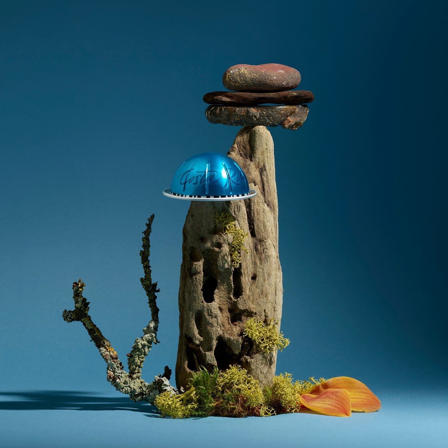 Still life shot of an aluminum coffee pod balanced on a wooden pedestal with rocks and moss against a blue background.