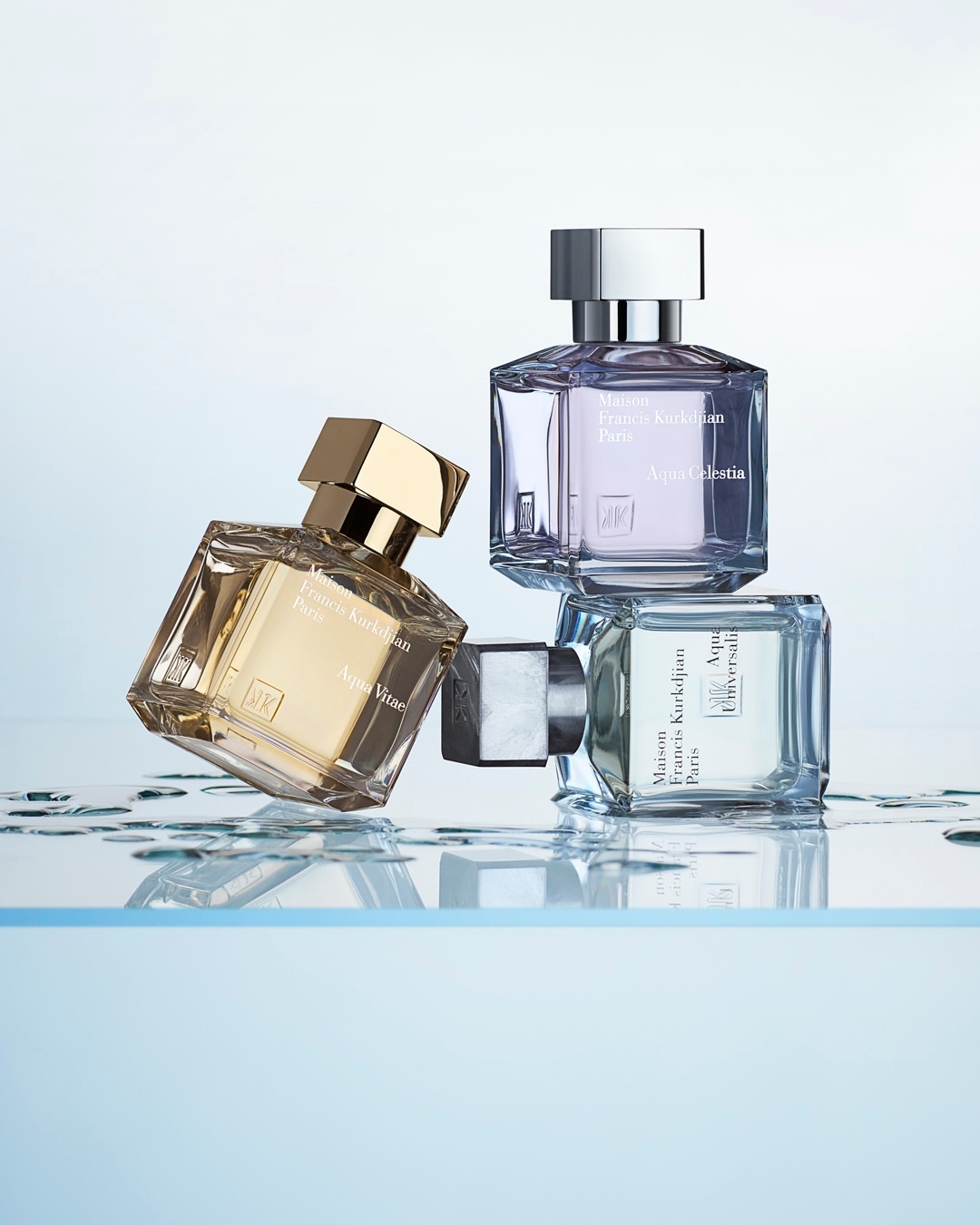 Three Maison Francis Kurkdjian perfume bottles are stacked on a clear surface, with a light blue gradient background.