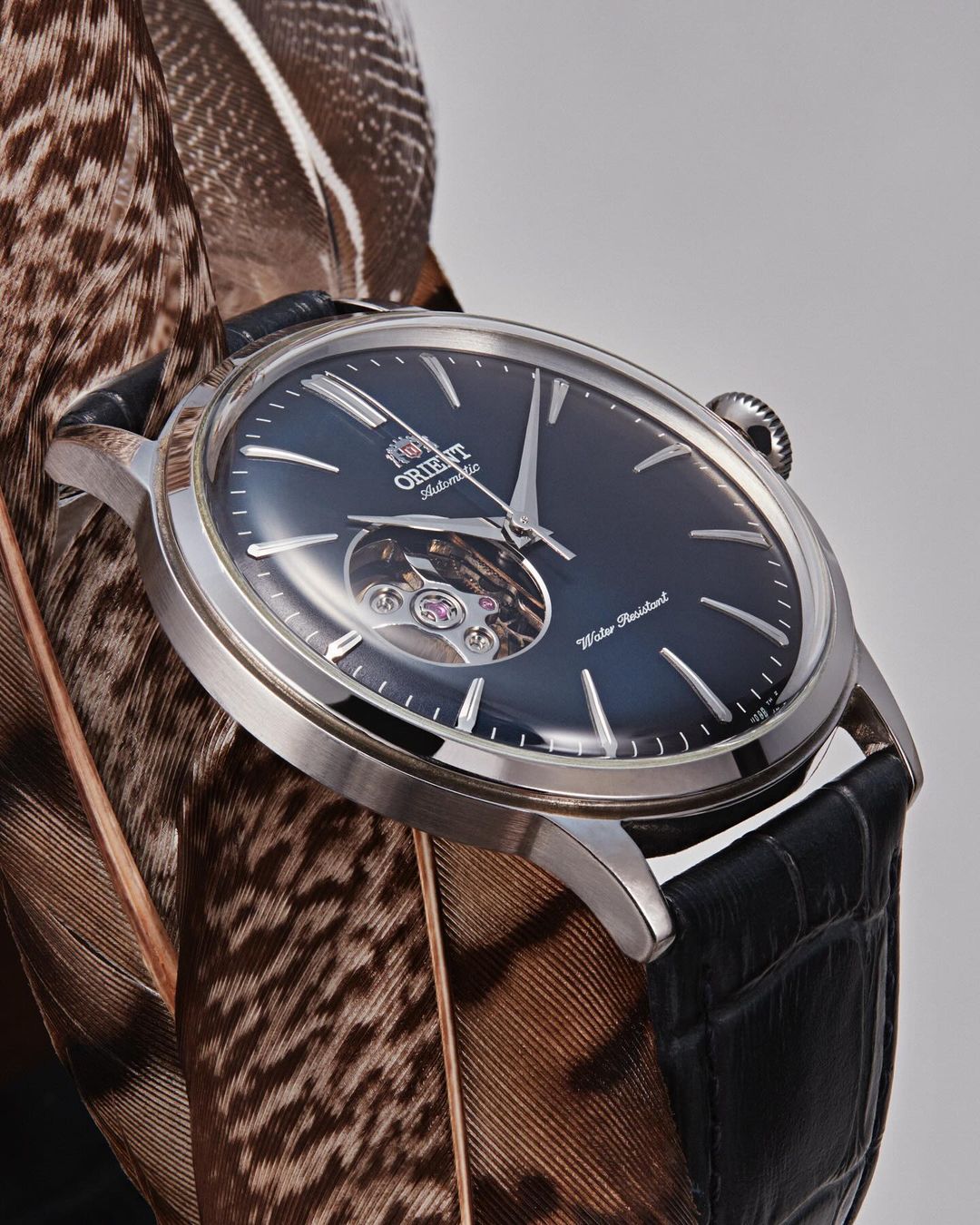 Orient automatic watch with a black band and silver casing sits atop brown and white feathers on a neutral background.