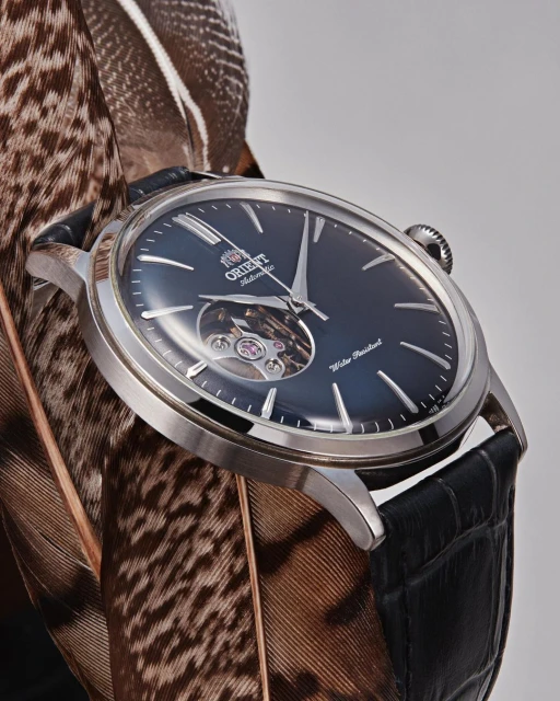 Orient automatic watch with a black band and silver casing sits atop brown and white feathers on a neutral background.