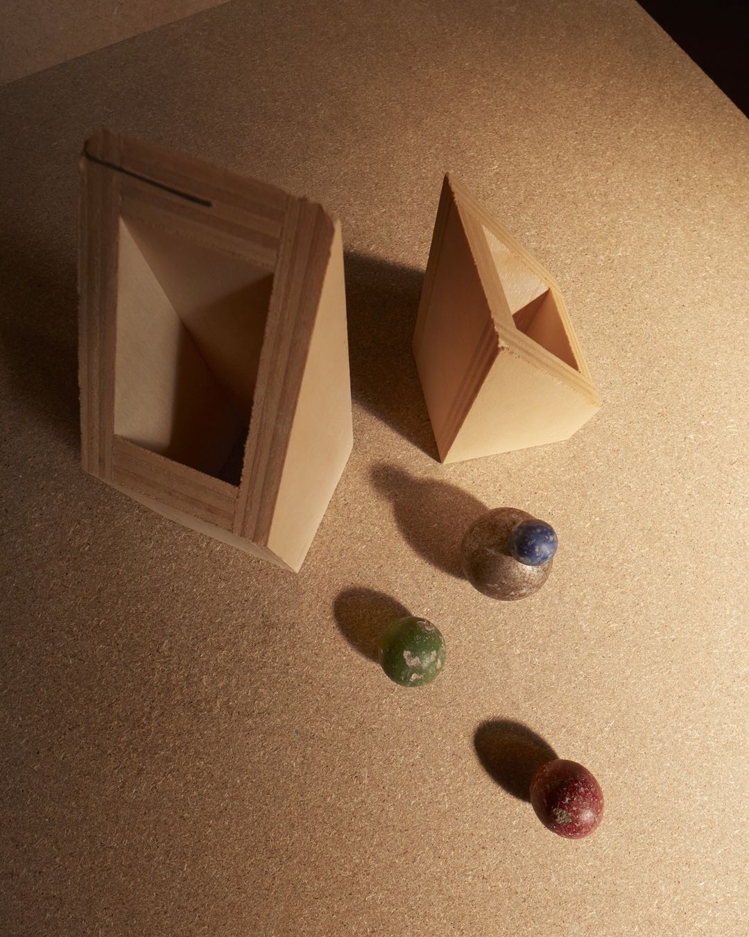 Two geometric, open, triangular wooden boxes arranged with three colorful marbles on a light brown surface, casting shadows.