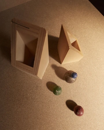 Two geometric, open, triangular wooden boxes arranged with three colorful marbles on a light brown surface, casting shadows.