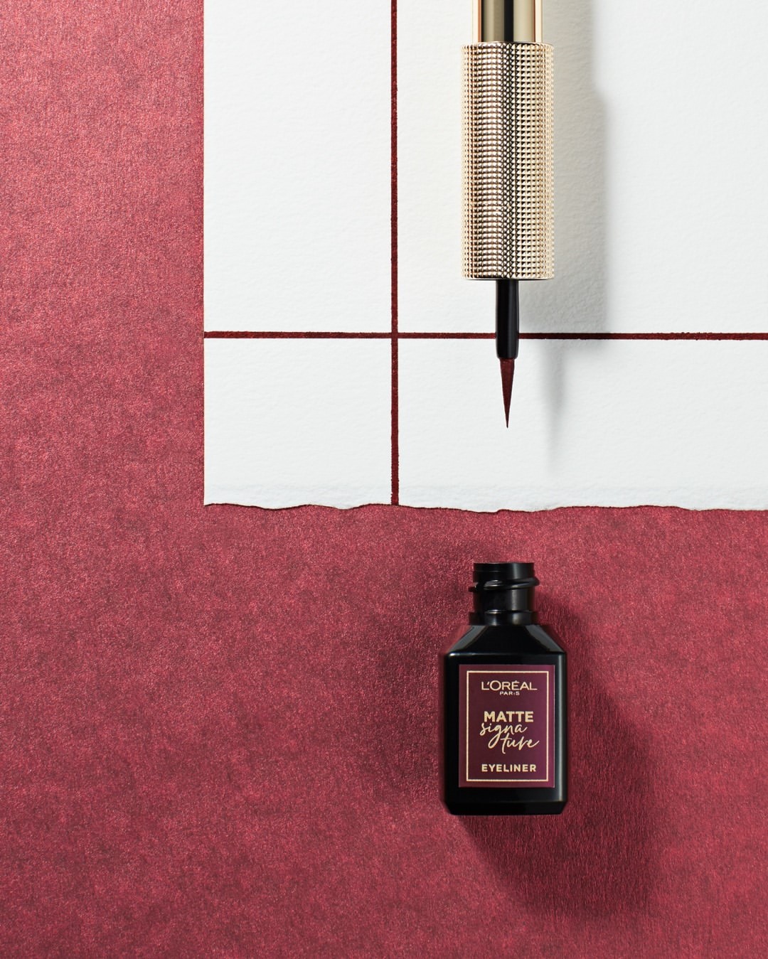 Close-up of L'Oreal Matte Signature Fuse Eyeliner product with wand applicator on white paper, against a textured burgundy background.