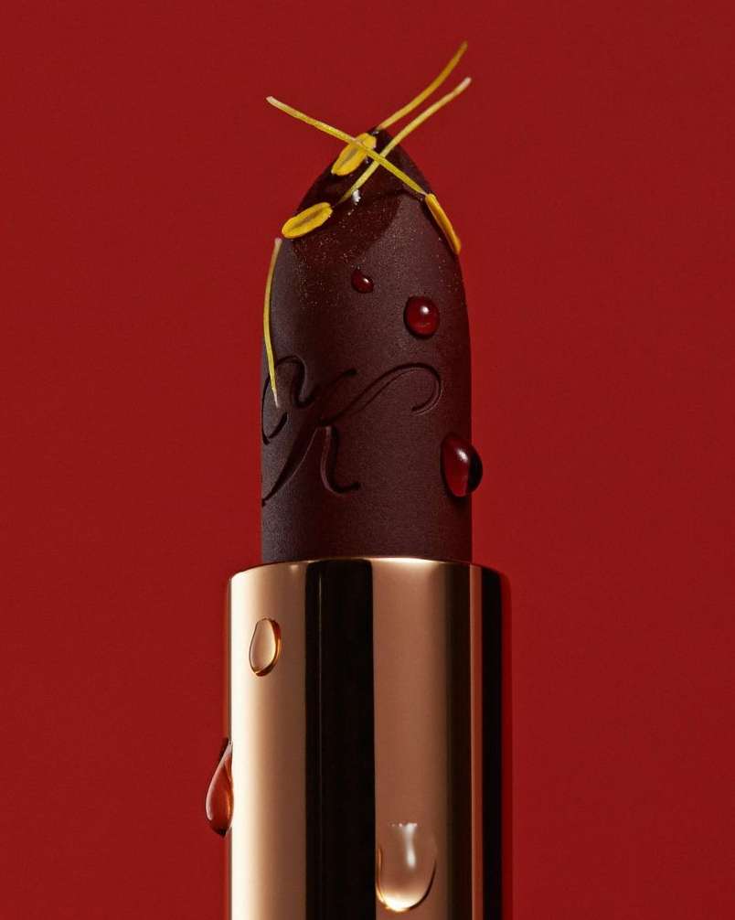 Close-up of a dark lipstick bullet with water droplets and yellow floral elements against a red background