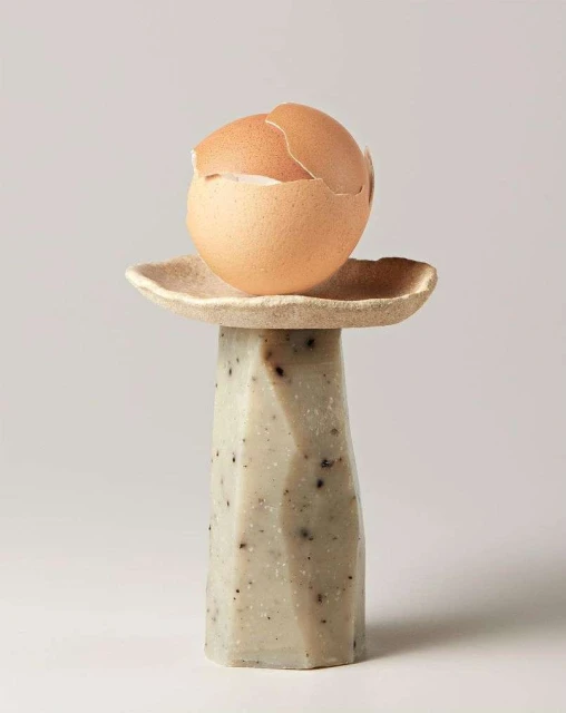 Still life shot of a cracked eggshell balanced on top of a geometric pedestal against a neutral background in a photo studio.