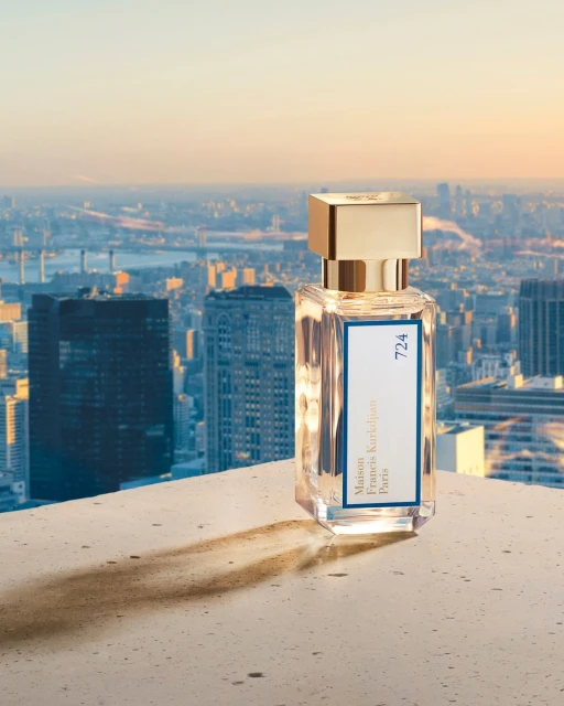 A bottle of Maison Francis Kurkdjian 724 perfume sits on a concrete surface with a cityscape and soft sunset in the background.