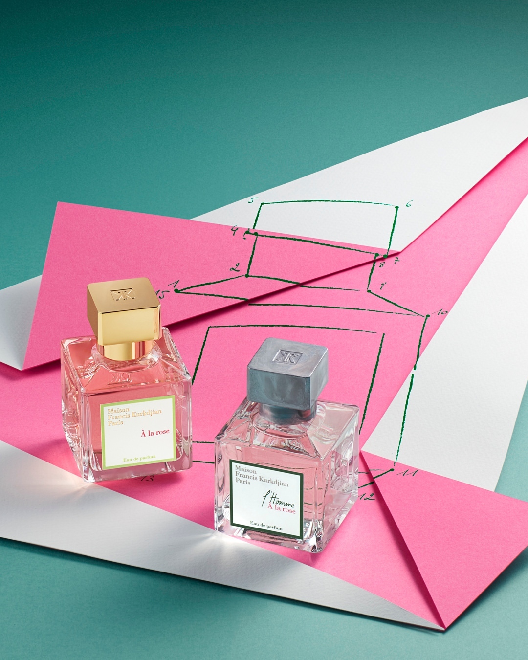 Two Maison Francis Kurkdjian perfume bottles stand on pink and white paper; fragrance product photography with teal background.