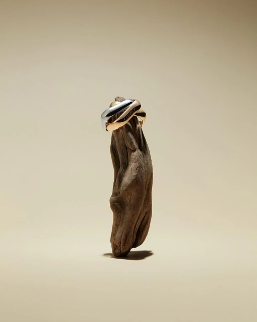 Three gold, rose gold, and white gold trinity rings are stacked on a textured brown tree branch against a neutral backdrop.