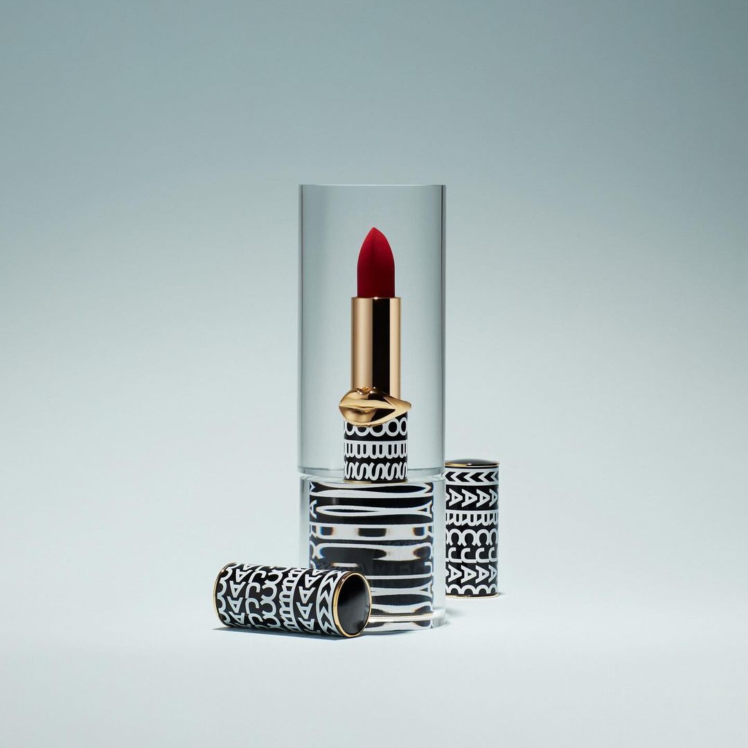 Close-up studio shot of red lipstick with patterned black and white case, displayed in a clear acrylic stand on a light grey background.