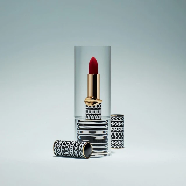 Close-up studio shot of red lipstick with patterned black and white case, displayed in a clear acrylic stand on a light grey background.