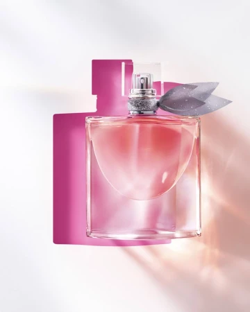 Square glass perfume bottle containing pink liquid with a grey fabric bow, set against white.