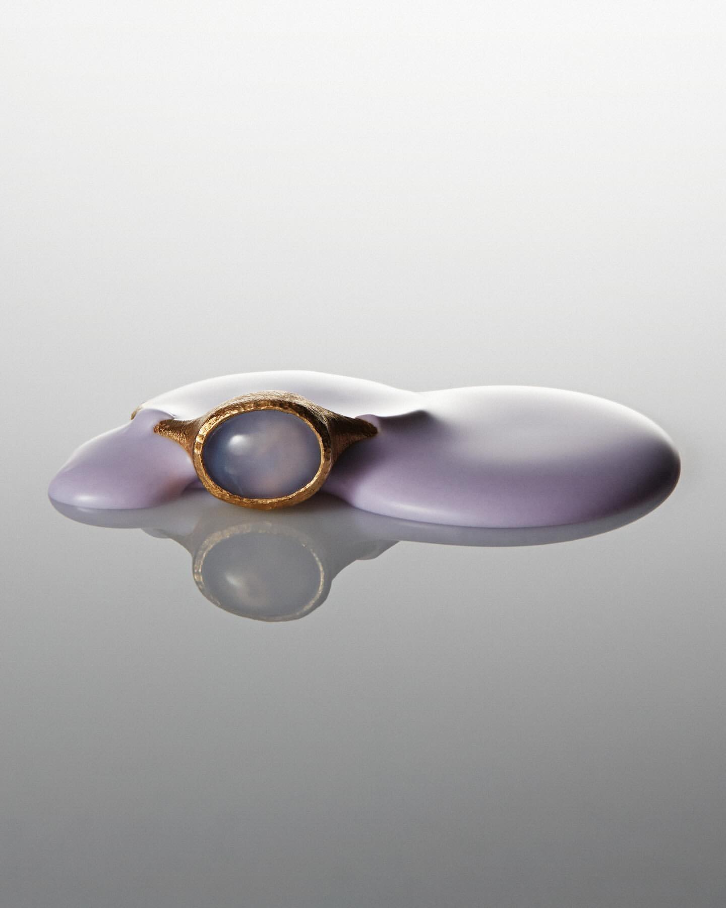 Close-up studio shot of a golden ring with a pale blue gemstone on a glossy lavender surface, set against a gradated grey backdrop.