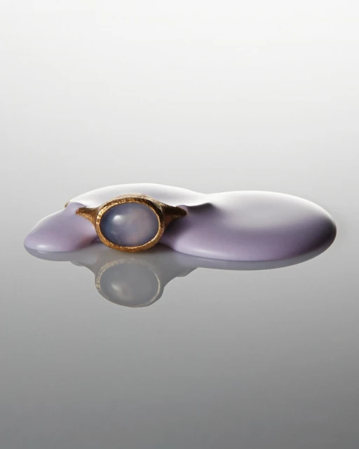 Close-up studio shot of a golden ring with a pale blue gemstone on a glossy lavender surface, set against a gradated grey backdrop.