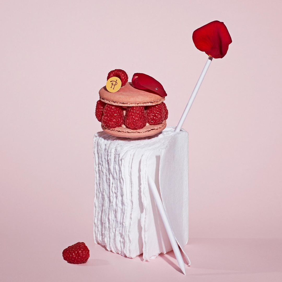Close-up of a raspberry macaron stacked on a white block with rose petal accents against a light pink background in a studio setting.