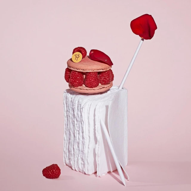 Close-up of a raspberry macaron stacked on a white block with rose petal accents against a light pink background in a studio setting.