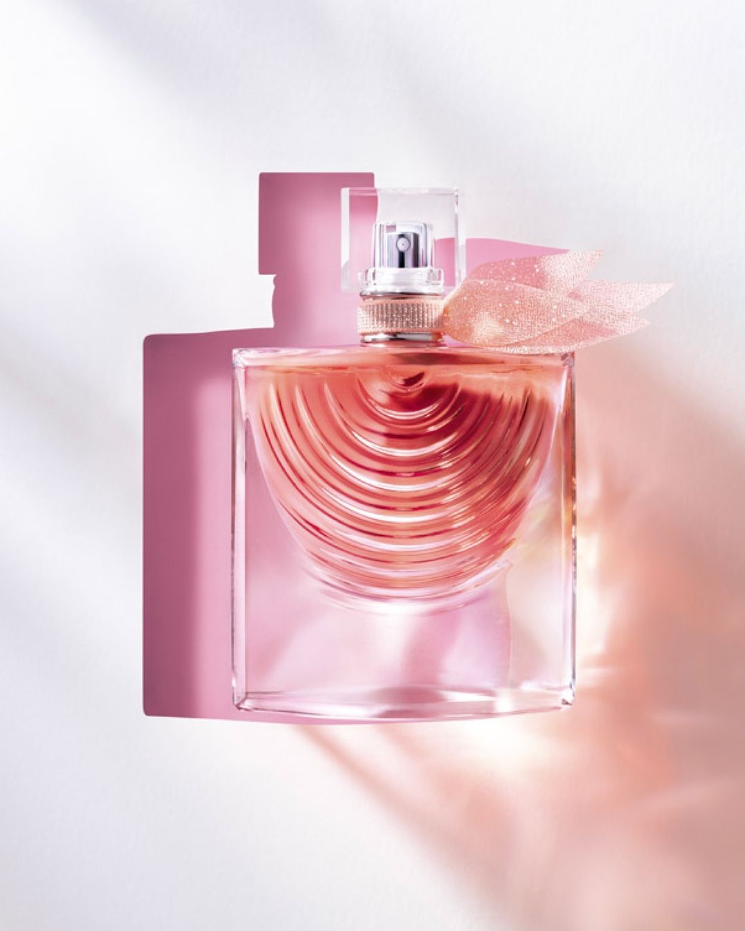 Overhead studio shot of a clear glass perfume bottle with a transparent lid and pink liquid on a white surface with pink shadows.