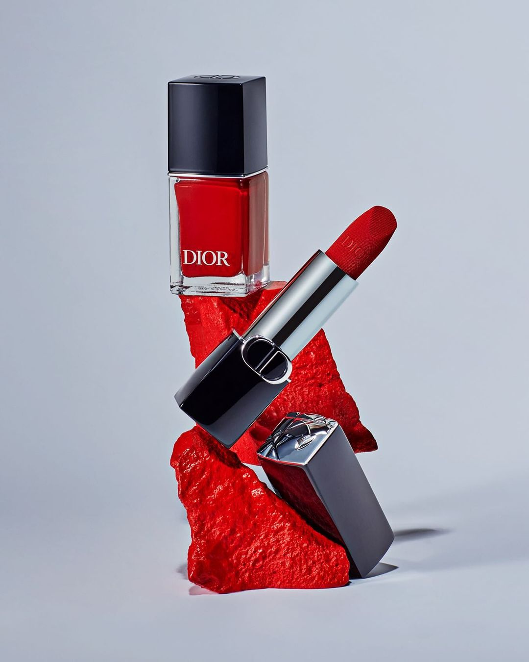 Still life shot of Dior nail polish and lipstick, stacked on red textured platforms, product photography in a light grey setting.