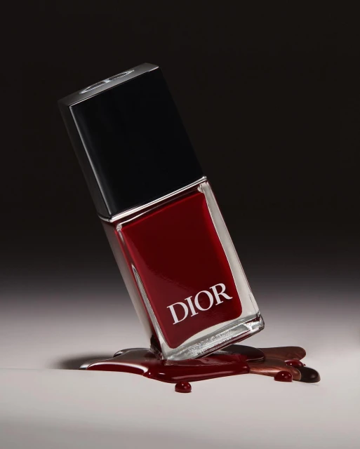Close-up of a leaning bottle of red Dior nail polish with the spilled contents forming a puddle on a light surface against a dark background.