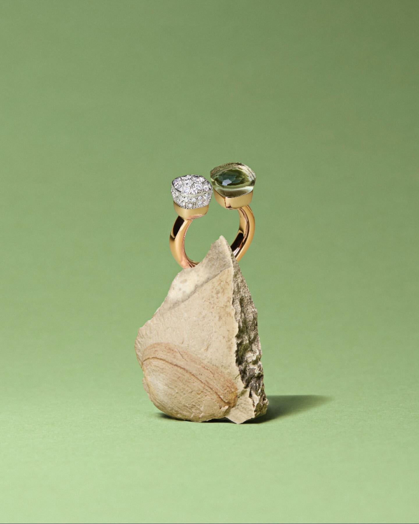 Gold ring with green and white gemstones displayed upright on a textured stone against a light green background, jewelry product shot.